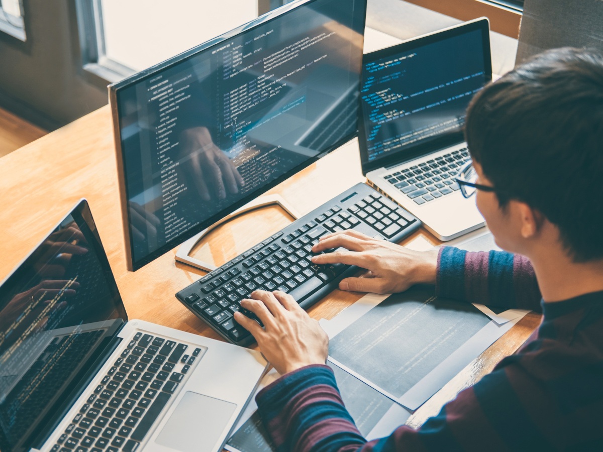 A man in glasses works on a coding project across multiple monitors and computers.