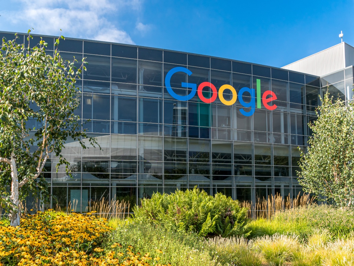 The Google logo on a large glass building, surrounded by trees.