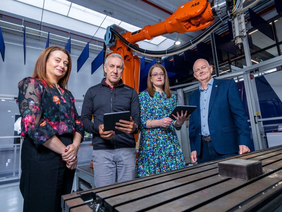 Four people standing in a sterile-looking research facility with manufacturing equipment behind them.