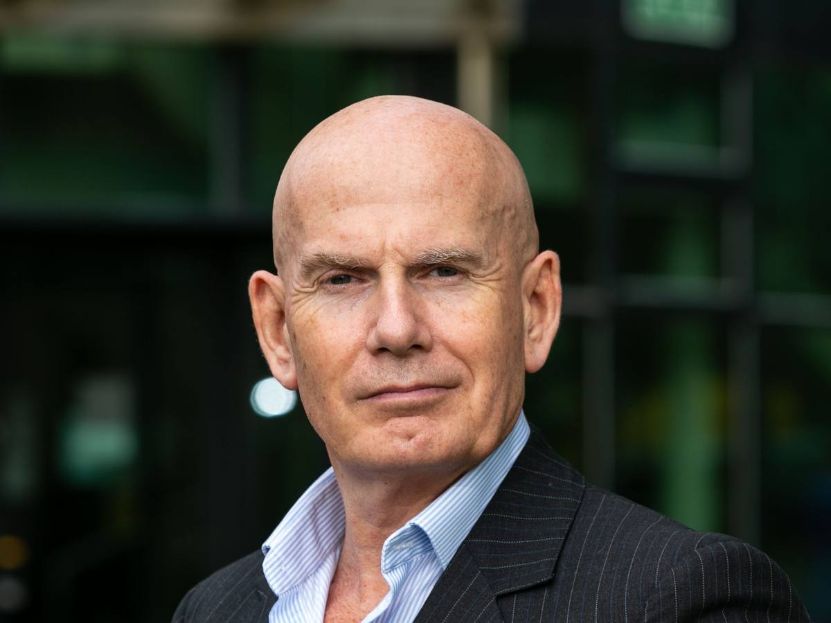 A bald man wearing a suit and open shirt collar with no tie stands looking at the camera.