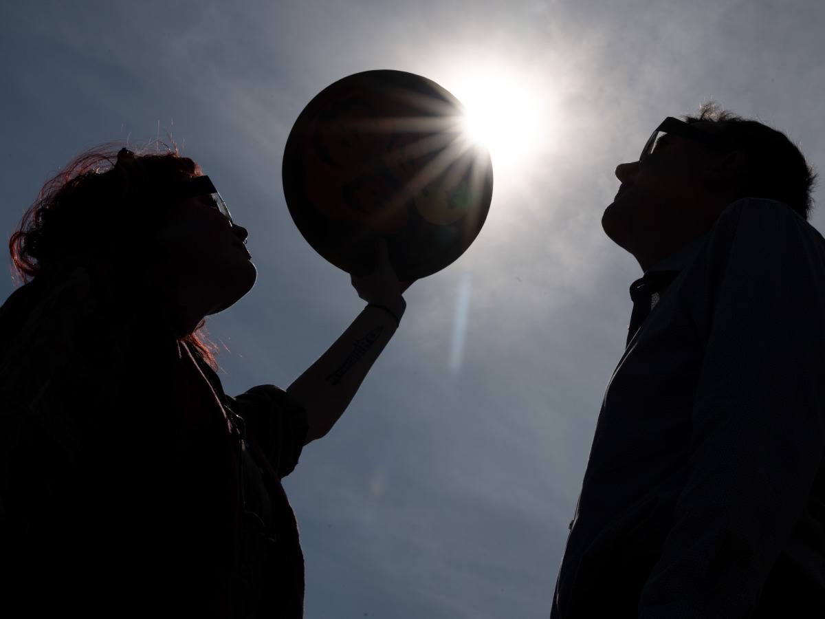 Silhouettes of two people with one person on the left holding up a circular object to block the sun, mimicking a solar eclipse.