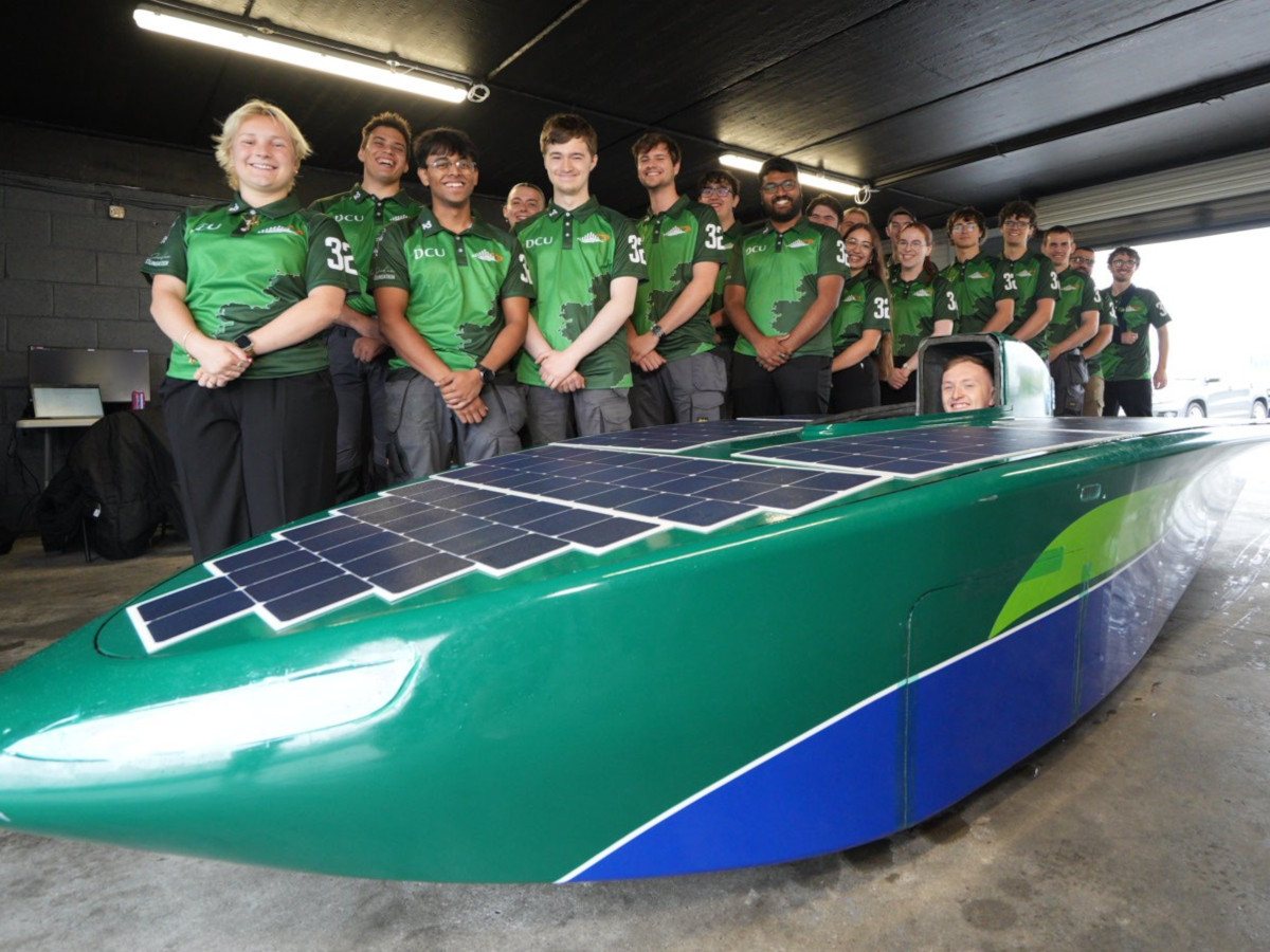 A group of students all wearing green DCU jerseys stand around a long green racing vehicle that