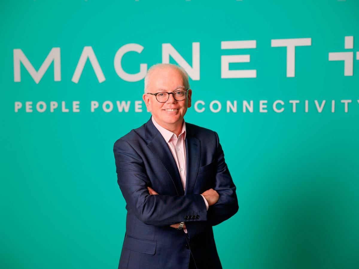 A picture of a man with white hair and glasses standing with his arms folded wearing a suit. He is standing in front of a teal coloured wall with the Magnet Plus logo on it.
