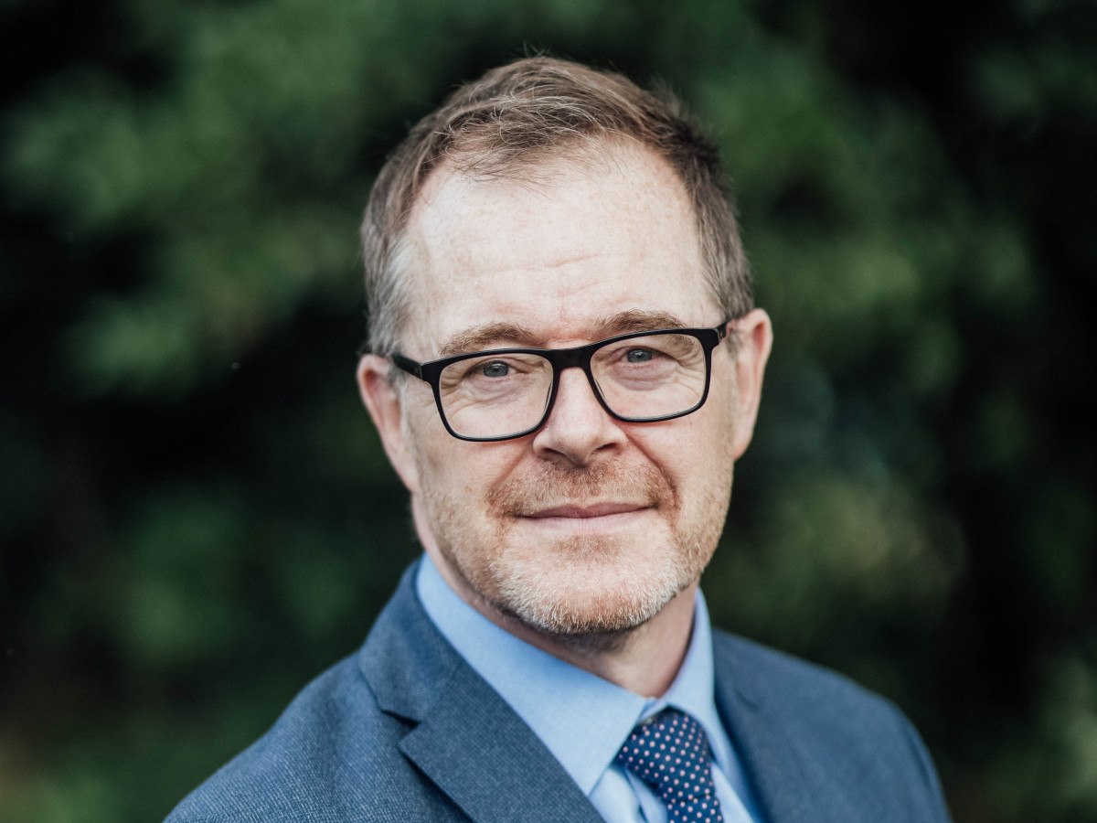 A picture of a bearded man wearing glasses and a suit and tie, standing in front of a nature background.
