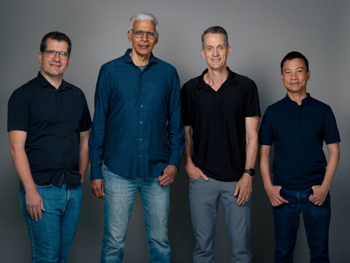 Four men in jeans and a shirt standing with their hands in their pockets.