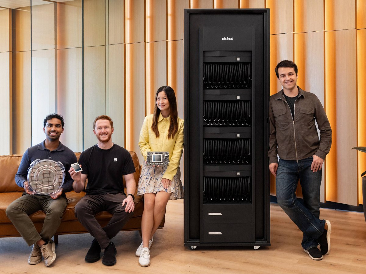 A picture of three men and one woman sitting on a couch and leaning against a data centre server rack in a brightly lit room with wooden furnishings.