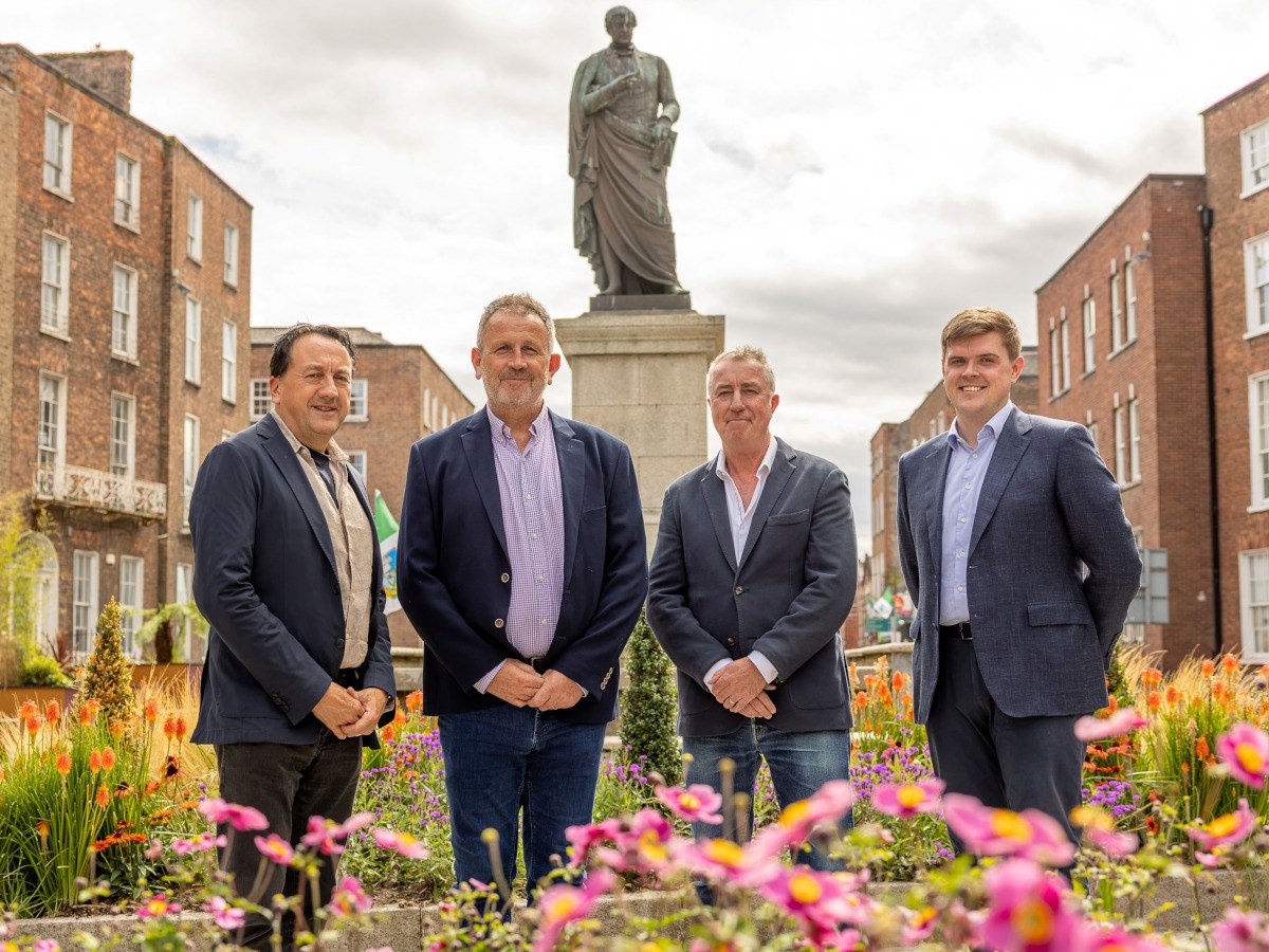 Four middle-aged men in business attire in front of a garden and large statue.