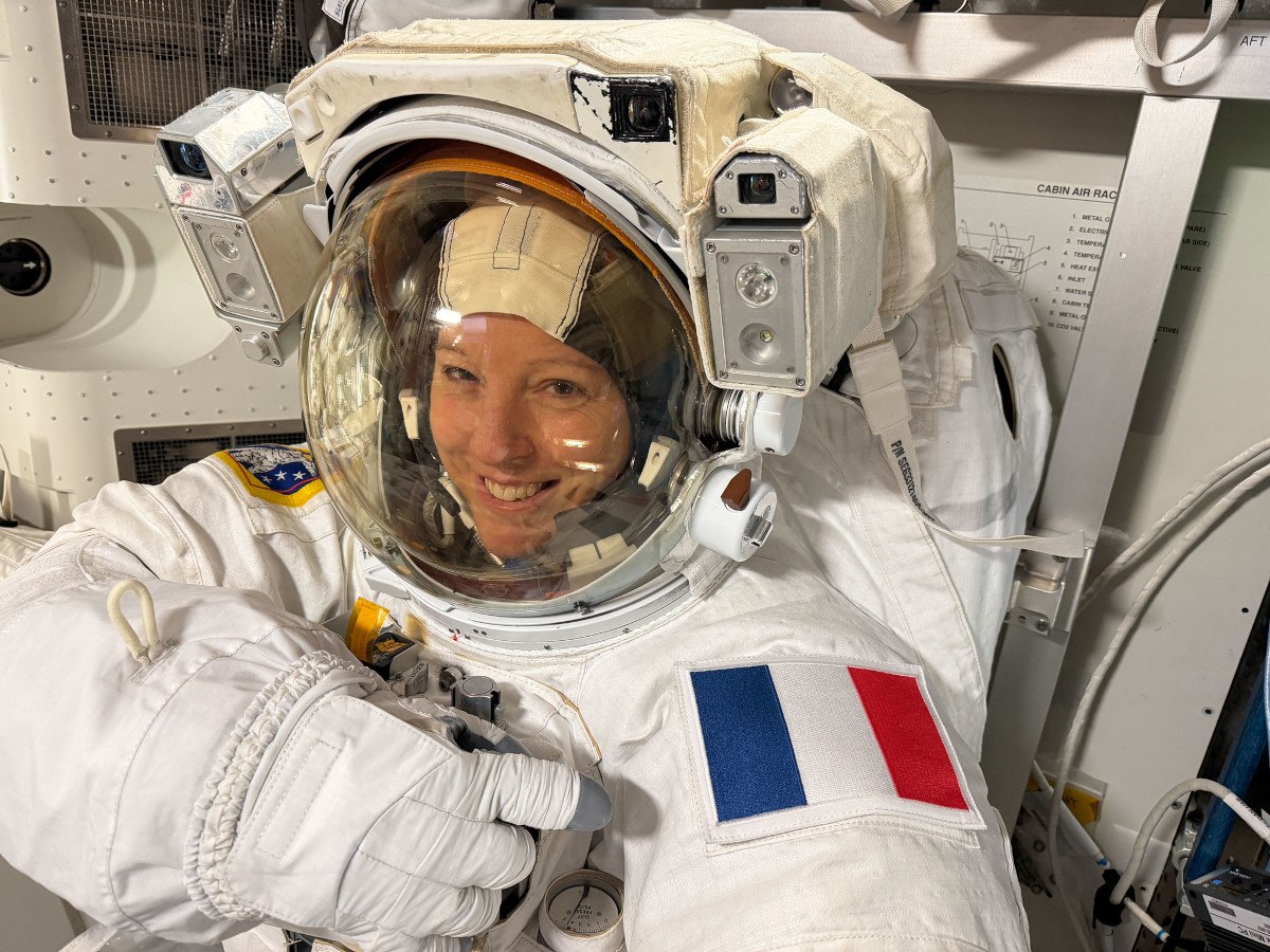 A woman wearing an ESA pressurised space suit smiles and points to her left shoulder, on which a patch of the French flag is displayed. She is Sophie Adenot.