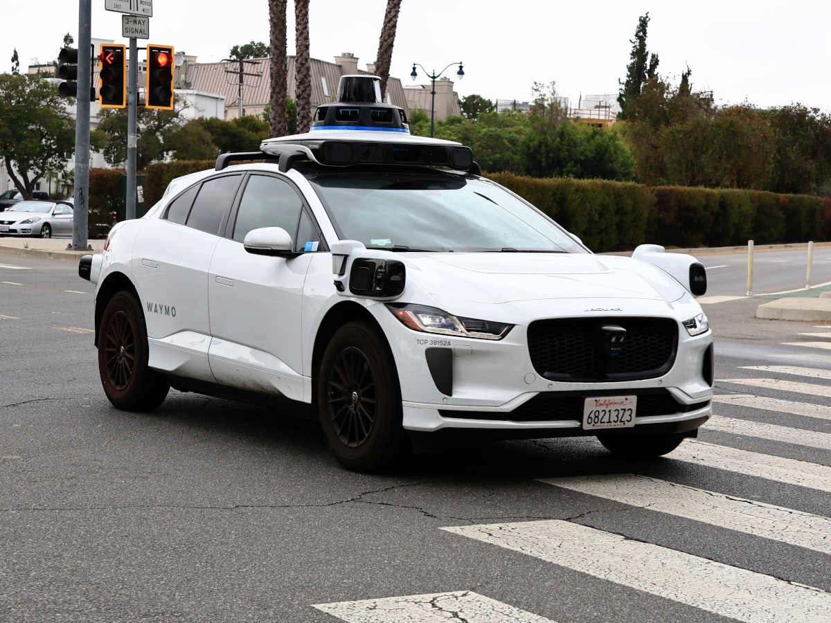 A white Waymo Jaguar I-Pace robotaxi operating on the a public street, driving through an intersection with orange traffic lights behind it.