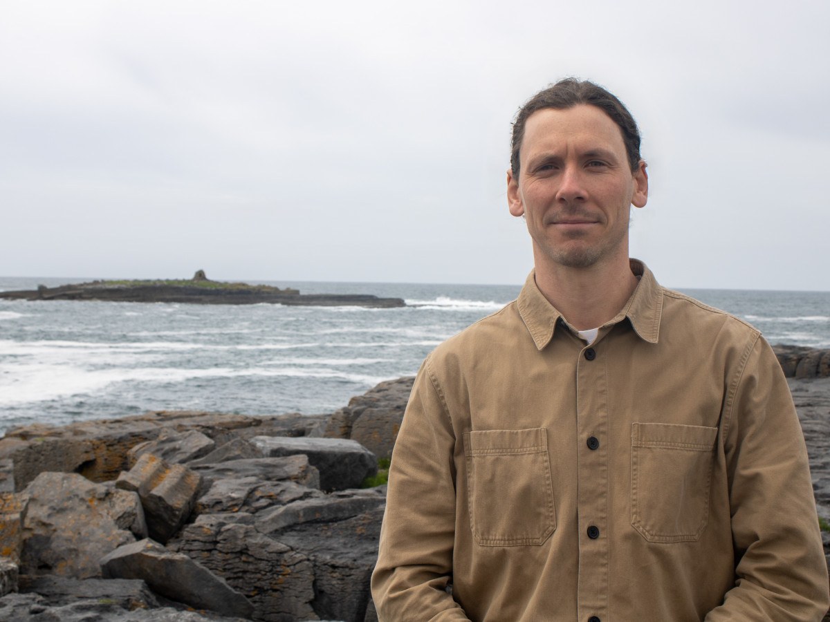 A man with brown hair and a brown shirt standing on a rocky coast with the sea in his background.
