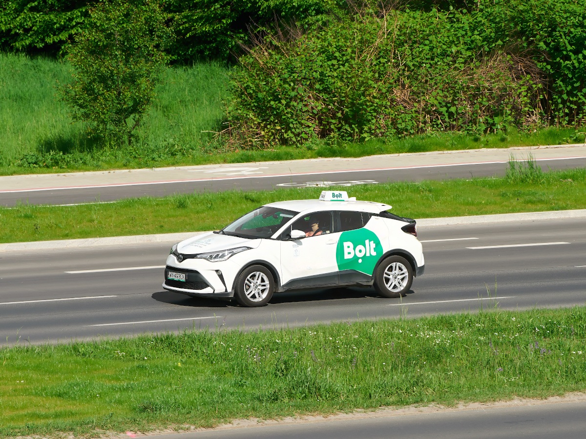 A white taxi with the green Bolt logo on the side drives along a road surrounded by greenery.