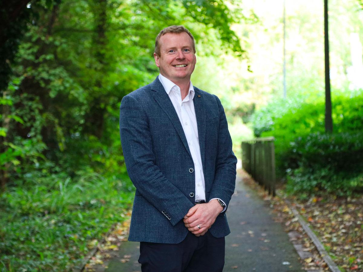 A man wearing a suit with a white shirt and no tie smiles with his hands clasped in front of him while standing in a woodland path.