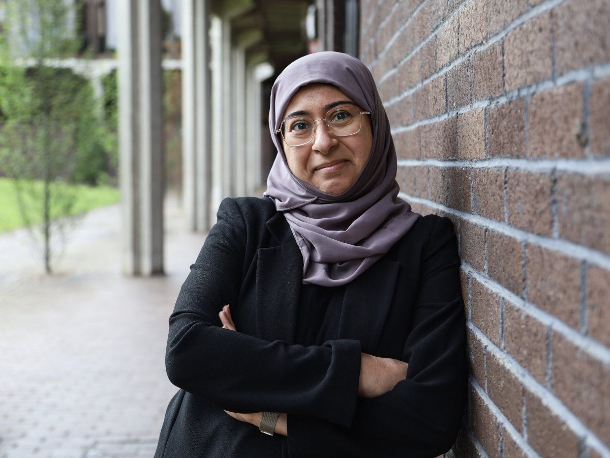 A woman wearing a hijab and glasses smiles while leaning against a brick wall with her arms folded.