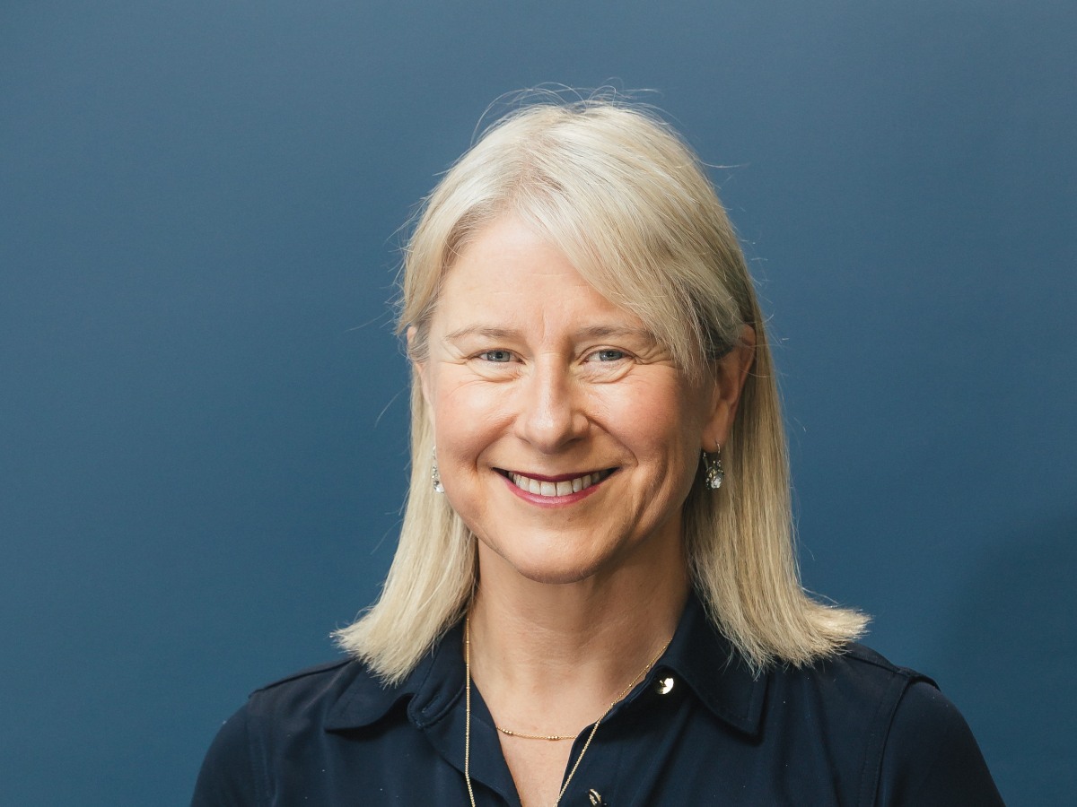 An older white woman with blond hair in a blue shirt, against a brighter blue wall.