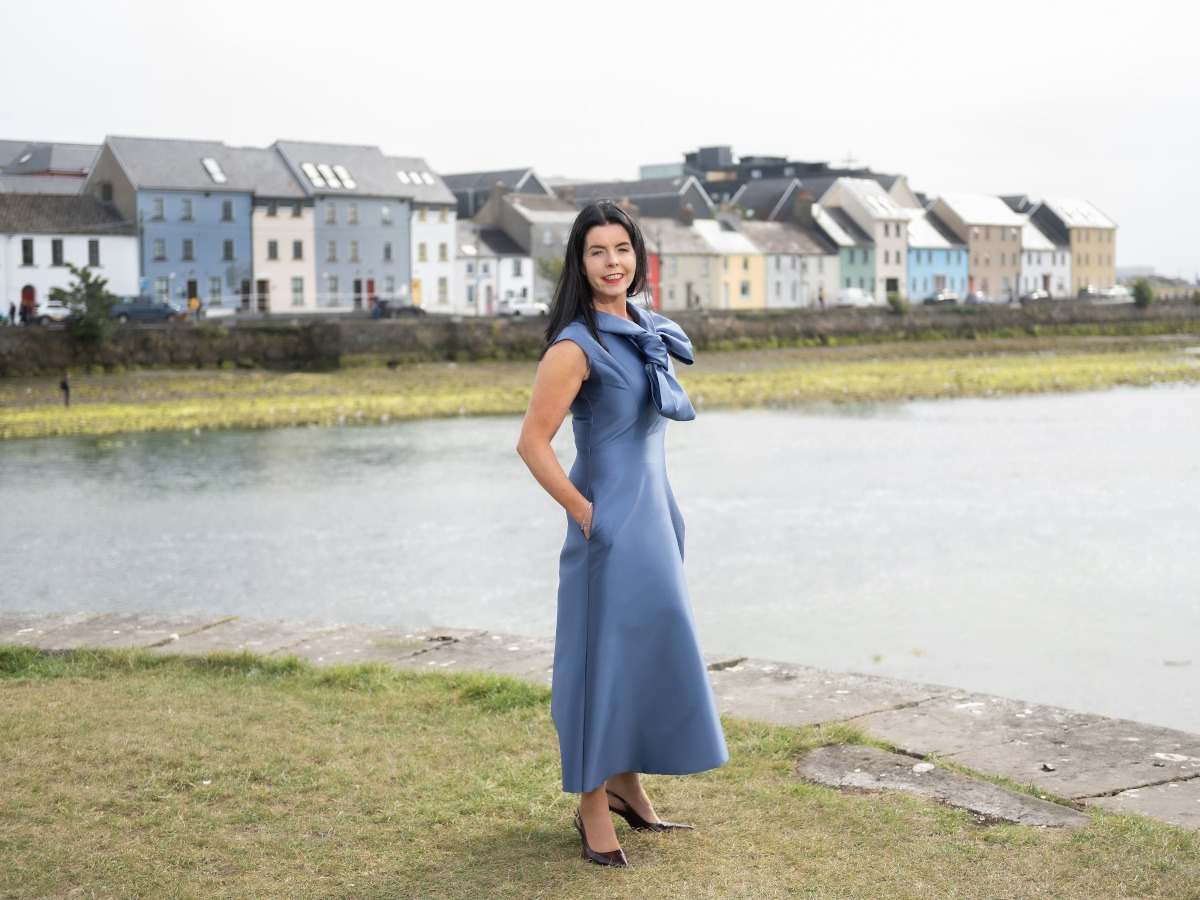 A middle aged white woman with long black hair, in a silk blue dress, in front of Galway