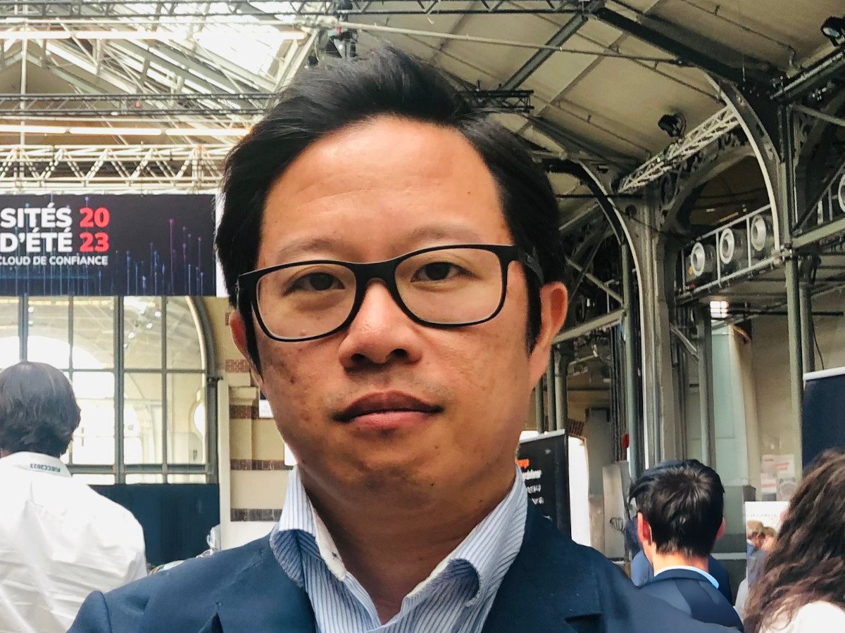 A picture of an Asian man with dark short hair and glasses wearing a navy suit with a light blue shirt with an open collar.