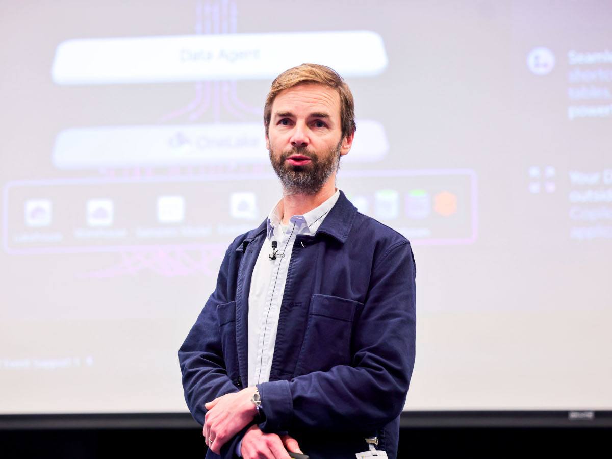 A bearded man wearing a suit jacket and white shirt with no tie stands up on a stage speaking with a browser home page projected on the screen behind him.