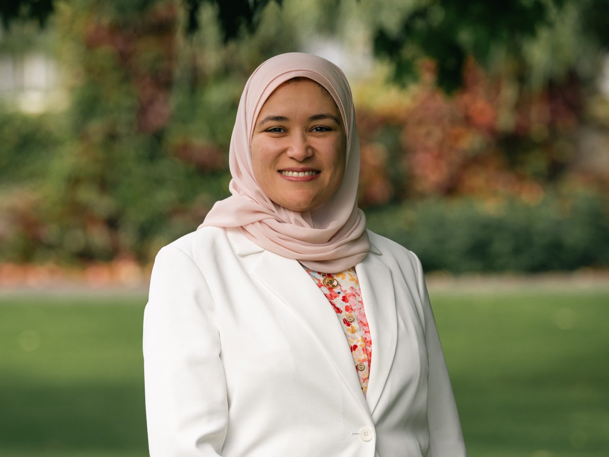 A young Middle Eastern woman with a beige headscarf in a white jacket in a garden setting.