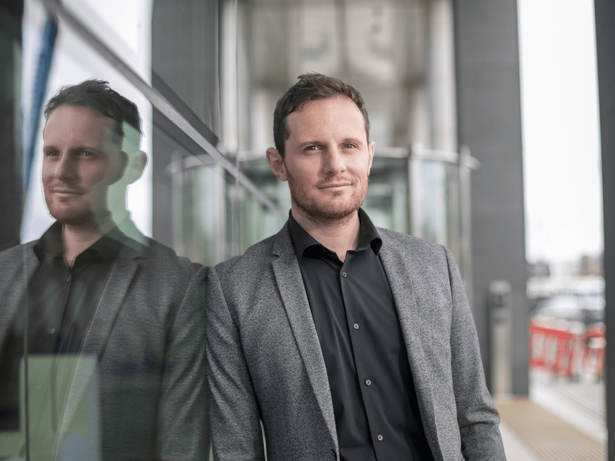 A young white fair haired man in a grey suit leaning against a glass building.