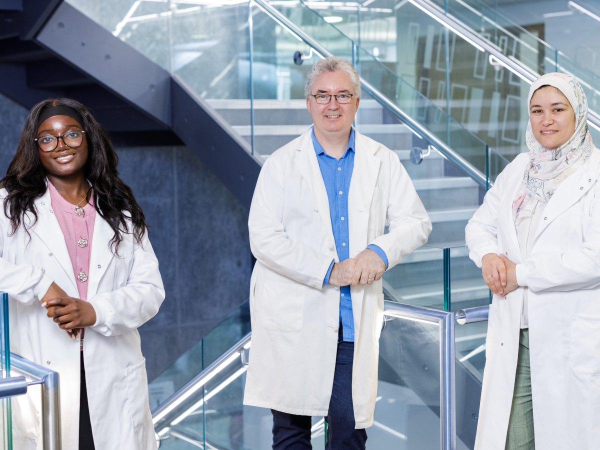 Two women and a man, diverse in age and ethnicity, pose in lab coats and smile in front of a metal and glass staircase.