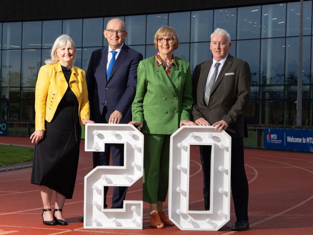 Four middle aged white people, gender diverse, in colourful and muted business attire, in front of a large 20 sign.