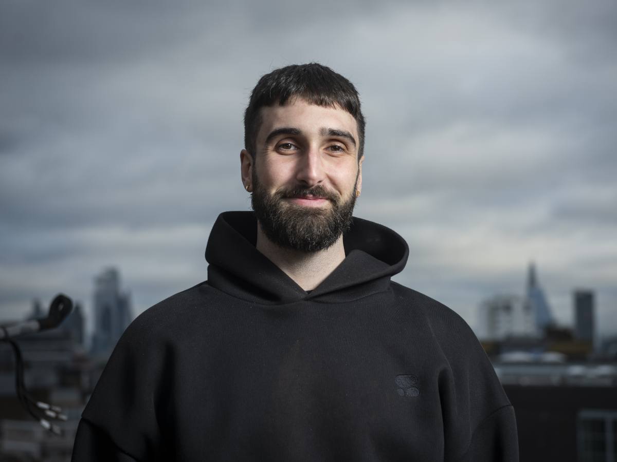A man with dark hair and a dark beard wearing a dark hoodie, in front of a city skyline backdrop.