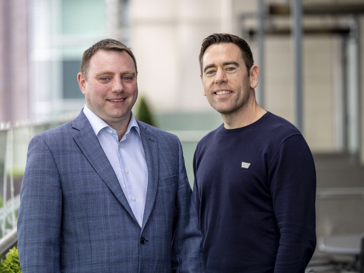 Two men standing together smile. One is wearing a grey suit and light shirt, the other is wearing a dark navy jumper.