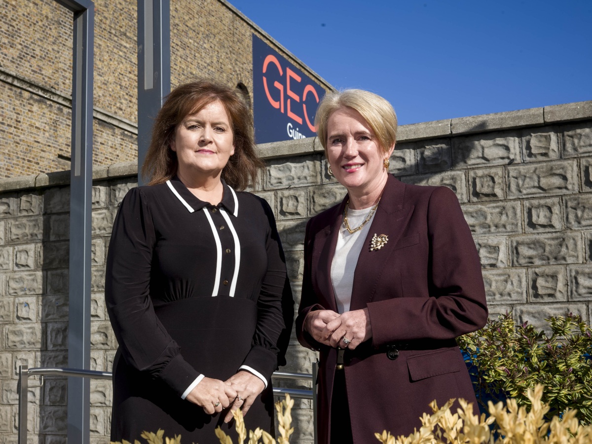 Two women wearing business attire stand together and smile in the sun.