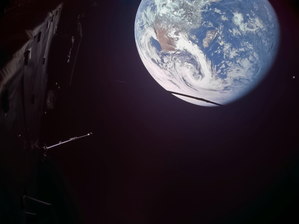 Mostly black space, most of Earth visible at the top right of the image. Part of a spacecraft visible on the left.