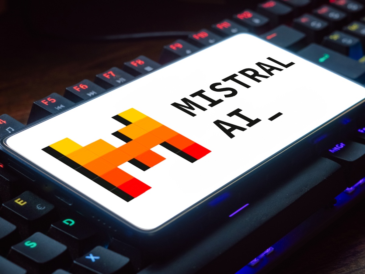 A Mistral AI logo on a white background on a phone screen held in front of an illuminated dark computer keyboard.
