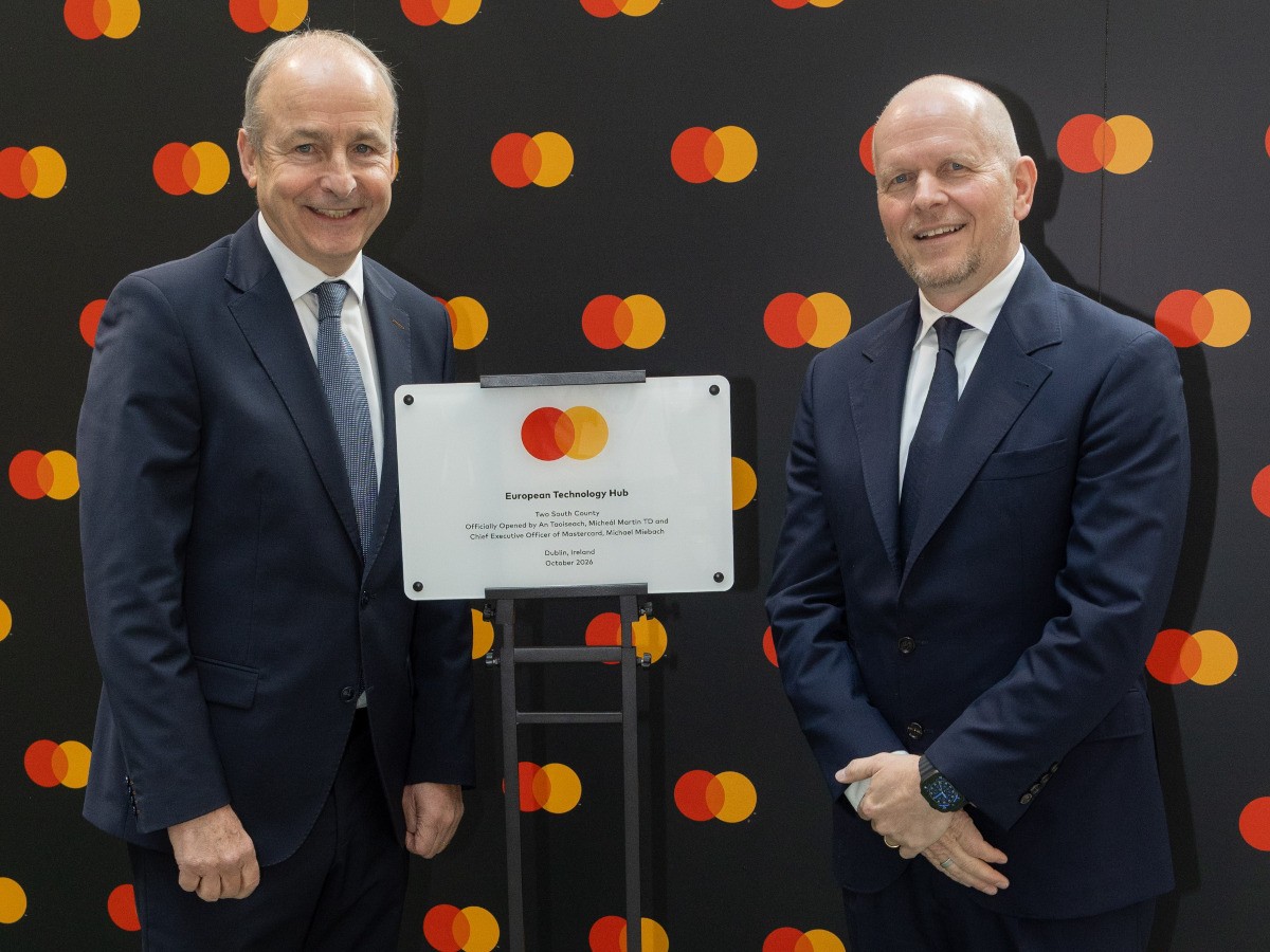 Two men in suits standing in front of a Mastercard banner.