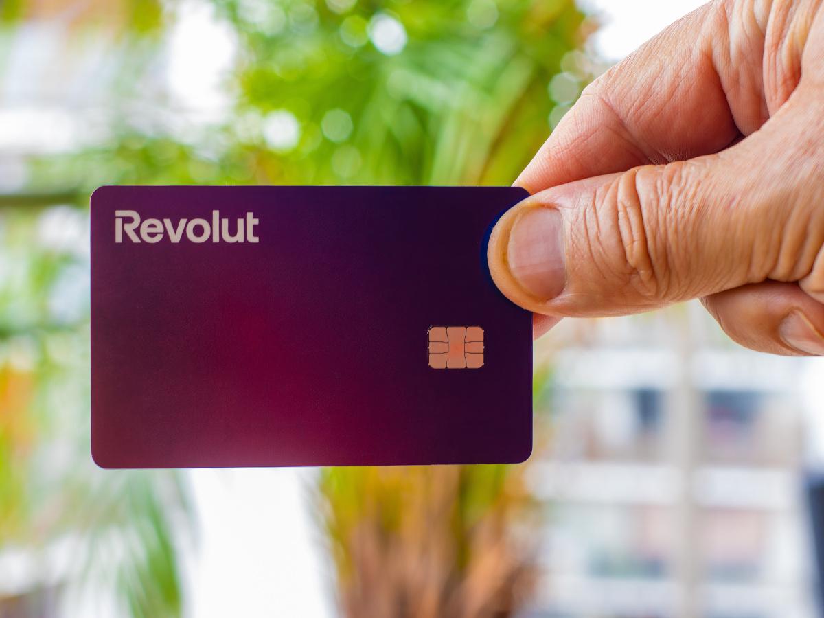 A purple Revolut bank card held high in a white hand, against green background.