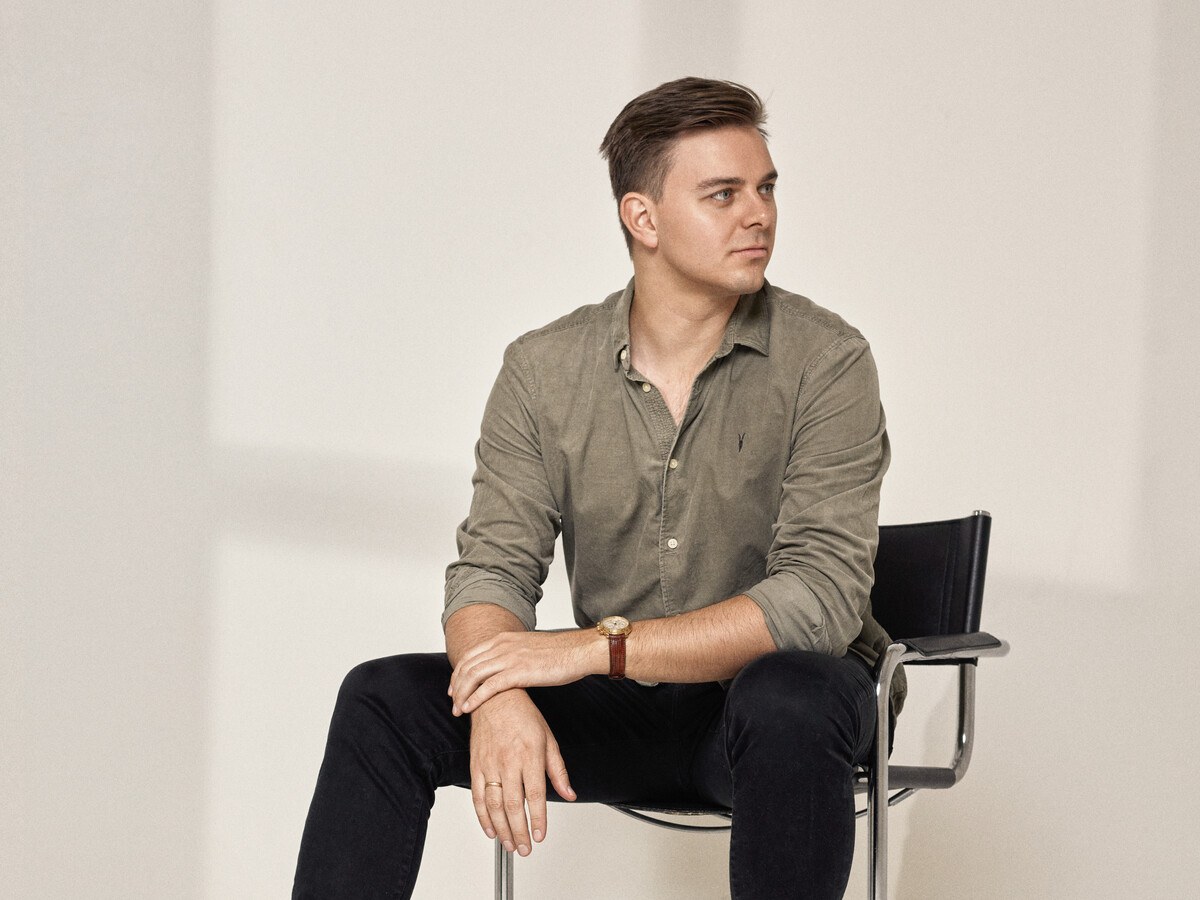 A man in a tan shirt, sitting on a chair, looking away.