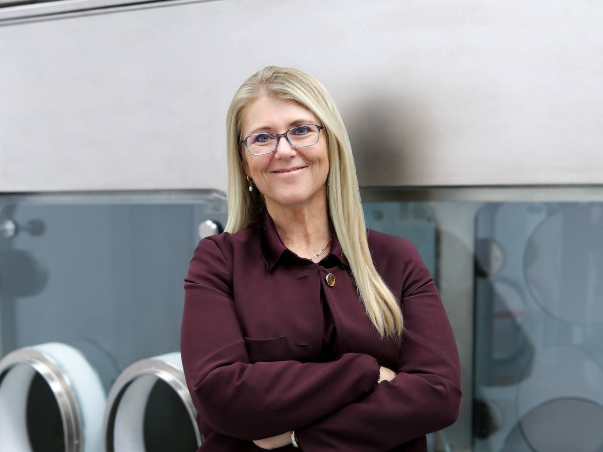 A middle-aged blonde woman in a maroon shirt and glasses leaning against cleanroom tech.