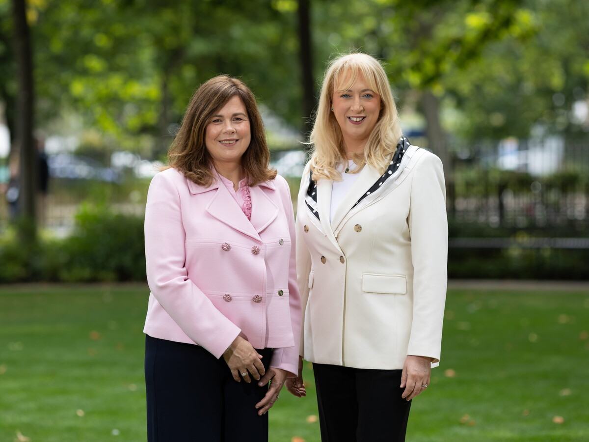 Two women in blazers standing in a park.