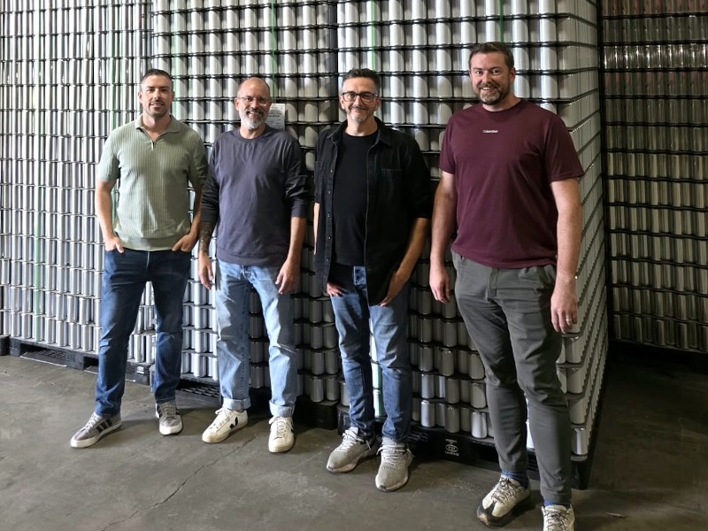 Four men stand in a line in front of stacks of drinks cans.